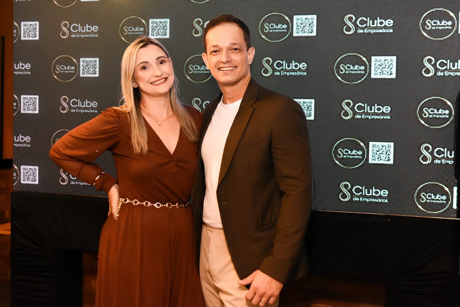 Dupla sorrindo diante do painel do Clube de Empresários